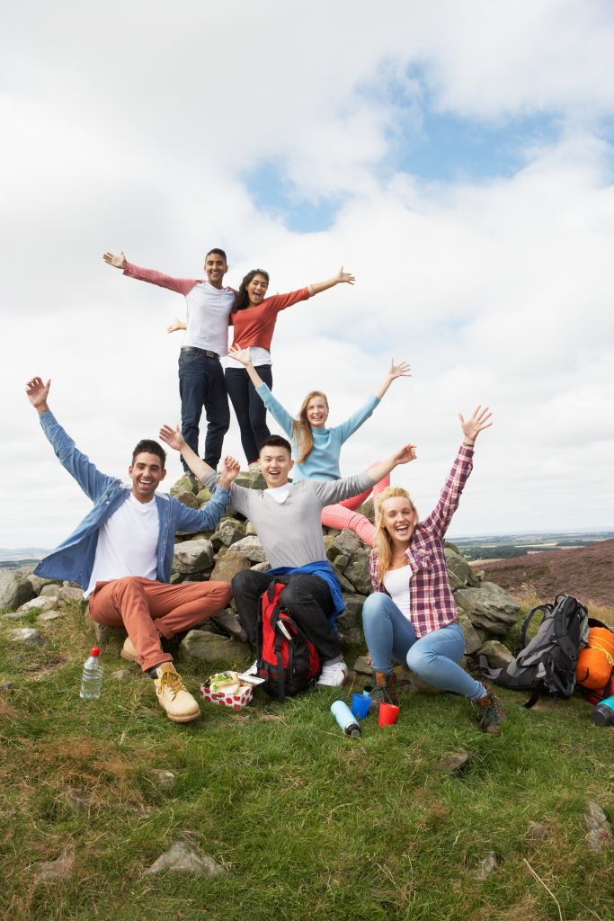 Picture shows a group of happy teenagers or young adults posing at the top of a hill. They are all excitedly raising their arms up to the sky.